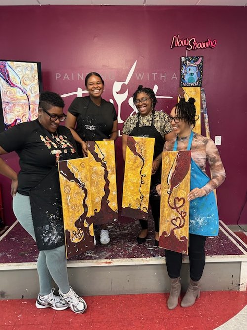 Four women at a painting class holding their completed abstract paintings. They are smiling and wearing apron smocks. The background shows a purple wall with art decorations and a sign that says 'Now Showing'.
