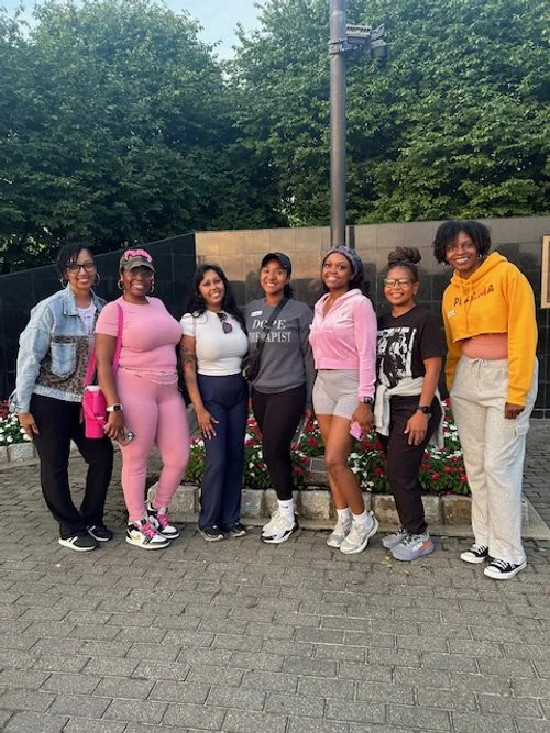 A group of seven young women standing together outdoors on a paved area, smiling, with trees and a lamp post in the background.
