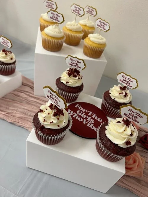 Cupcakes with white frosting and red sprinkles, labeled "For The Girls Who Rise," displayed on white boxes at a celebration event.