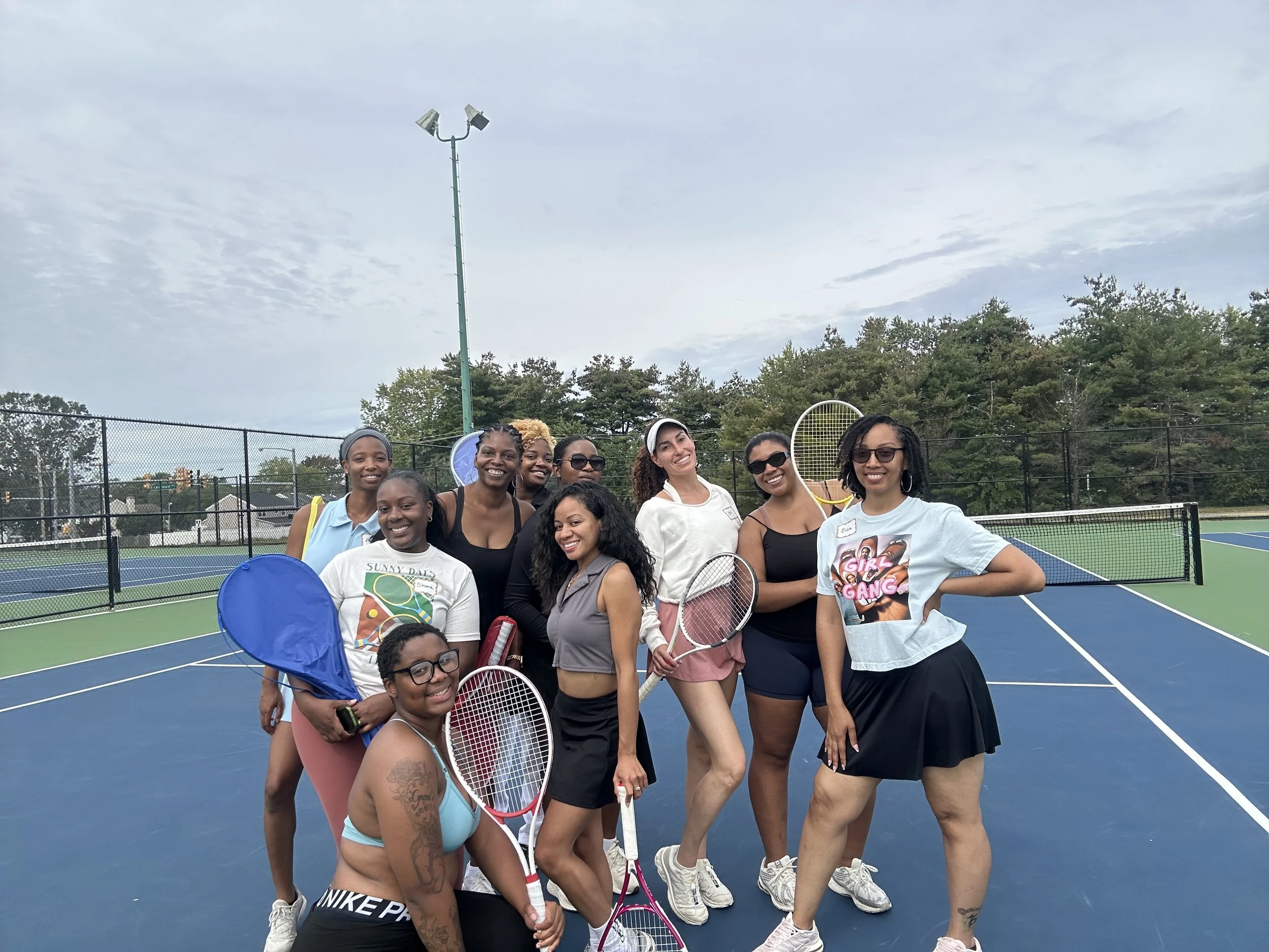 Group of nine women standing on a tennis court, holding tennis rackets, smiling, with trees and a cloudy sky in the background.