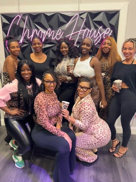 Group of nine women posing in front of a neon pink sign that reads 'Chrame House' with a black geometric wall background, some holding cups, dressed in various colorful and patterned outfits.