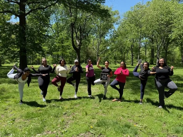 Eight women standing on grass in a park, posing with one leg lifted and holding various poses; some holding a guitar, a small chalkboard, and others with fists raised or hands on hips. Trees and a blue sky are in the background.