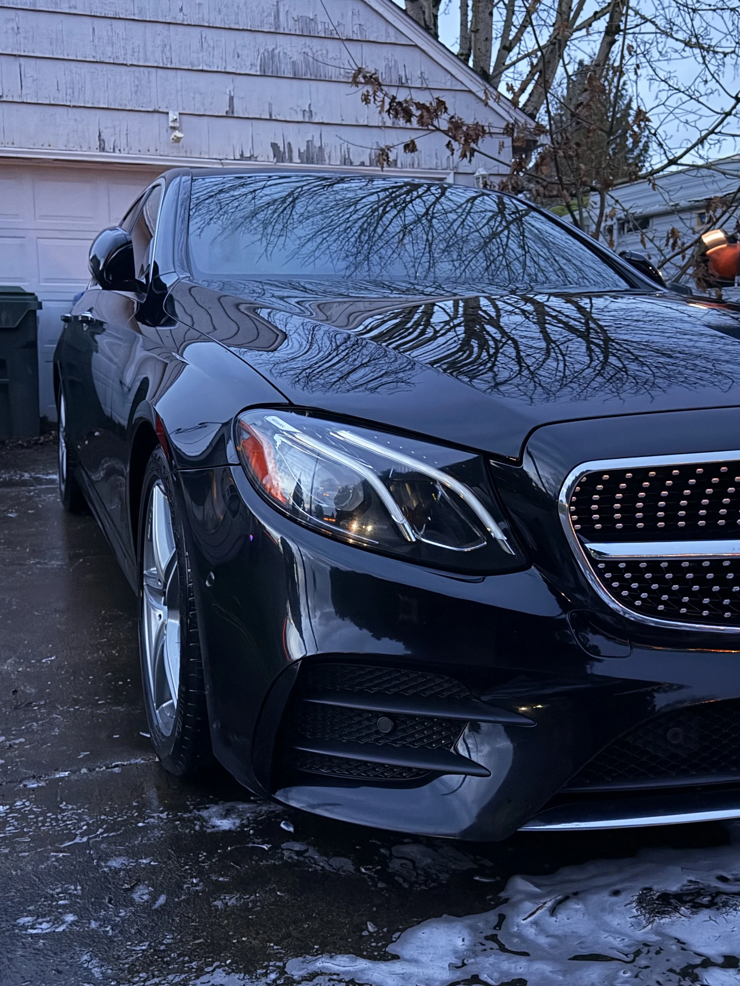 A black Mercedes-Benz parked in a driveway, with reflections of leafless trees on its surface.