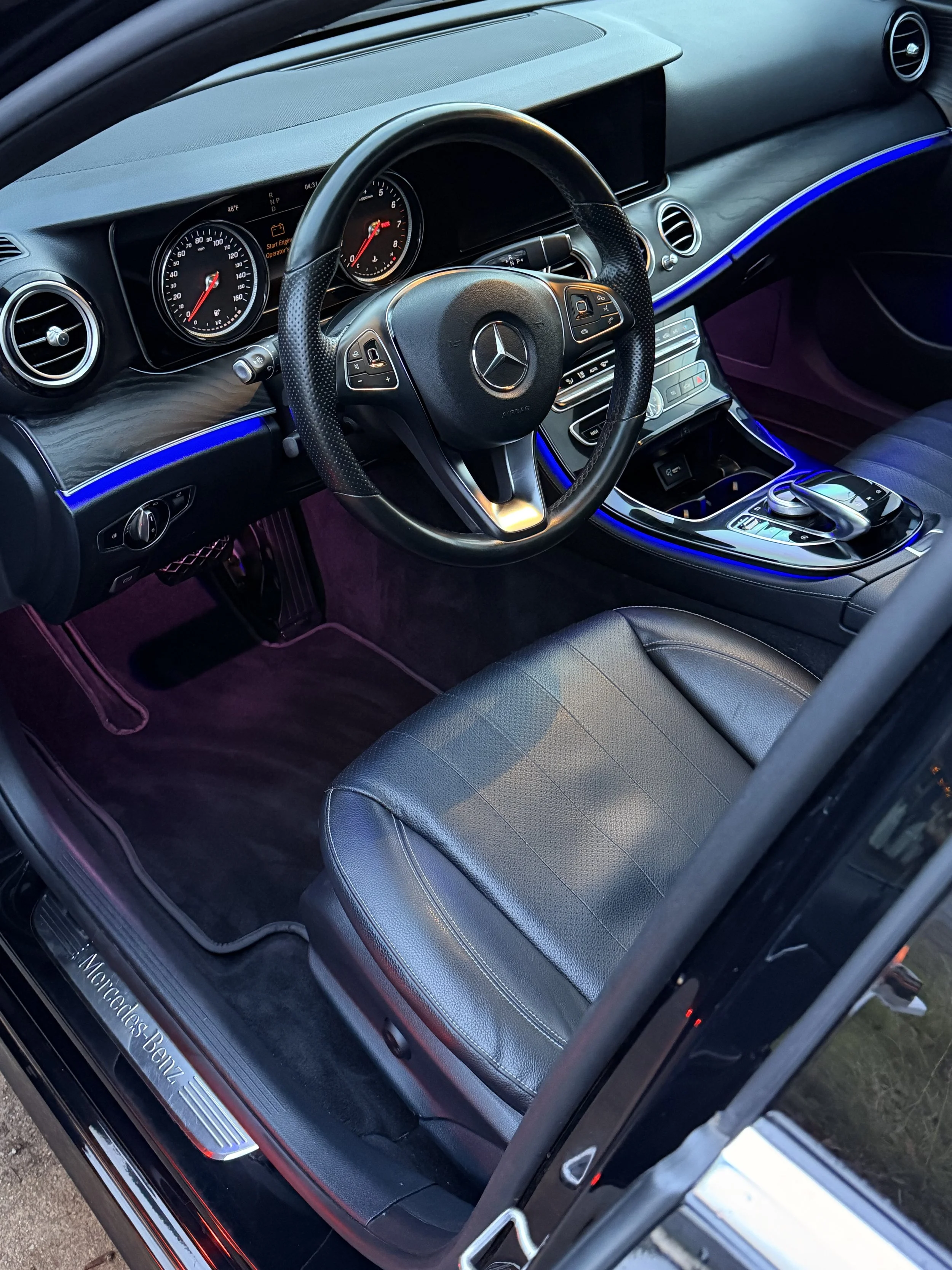 Interior of a black Mercedes-Benz car, showing the steering wheel, dashboard with instrument cluster, center console with controls, and leather seat with ambient lighting.