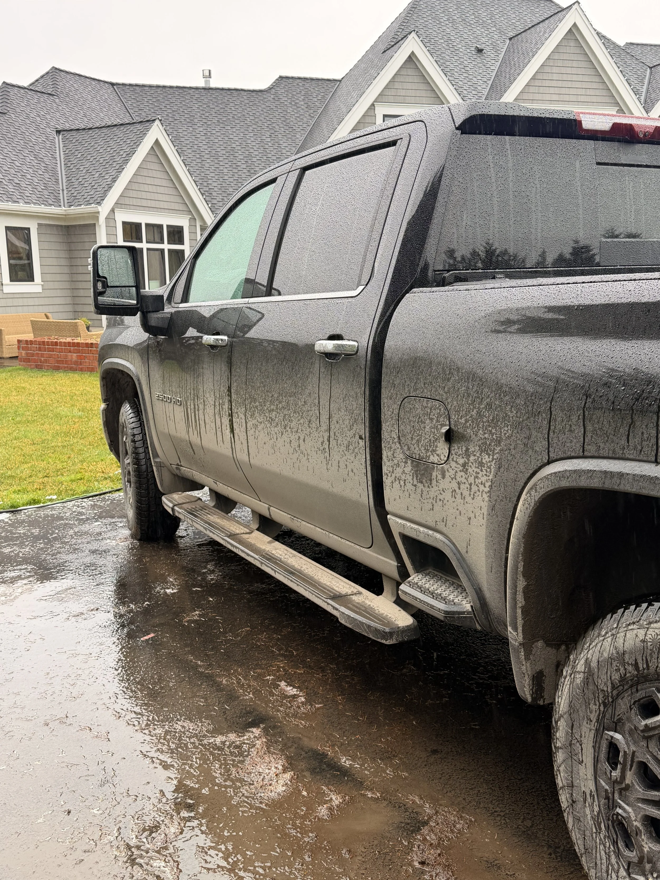 A gray pickup truck with water droplets on its surface parked on a wet driveway in front of a house with gray siding and a gabled roof.