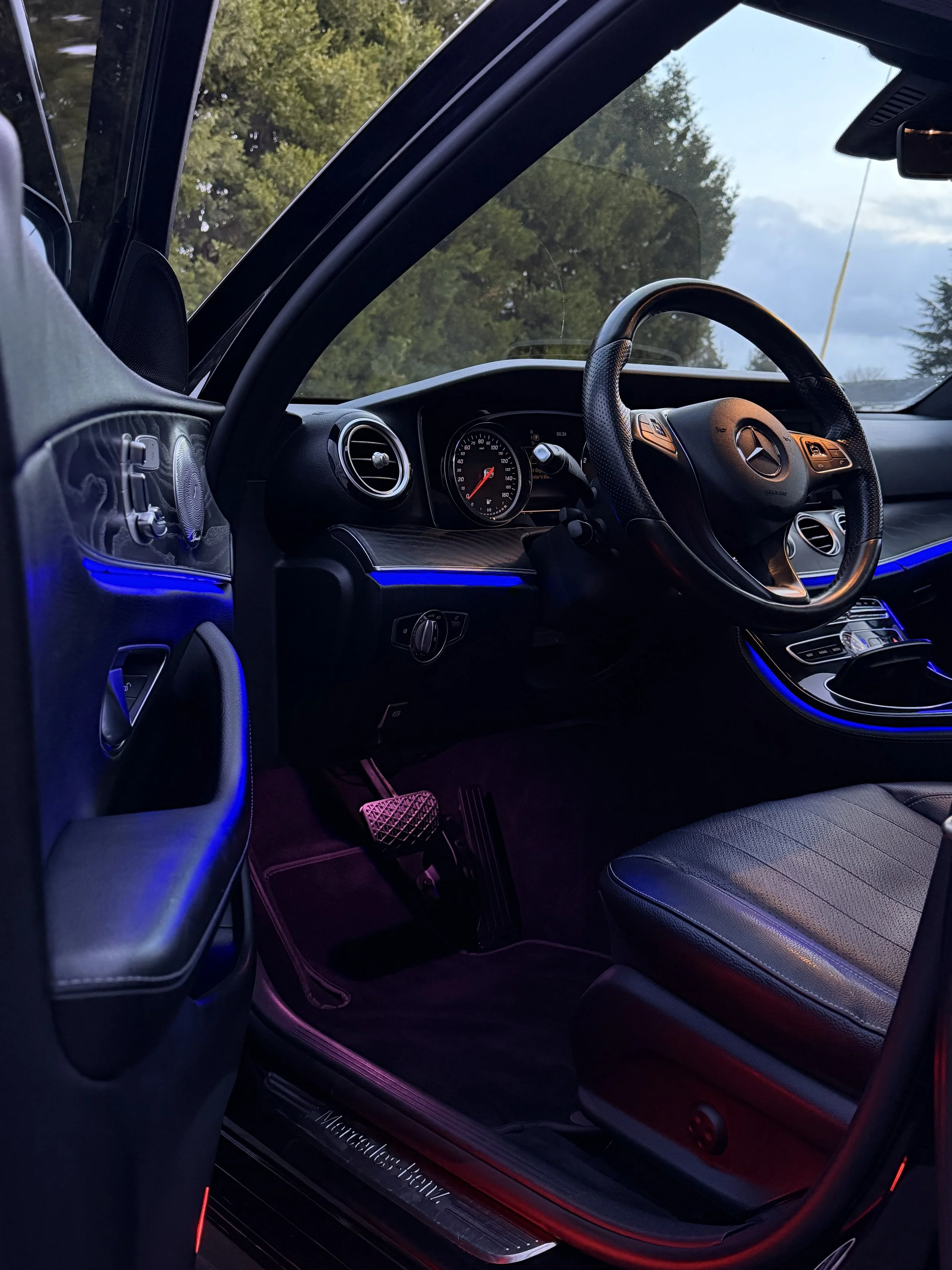 Interior of a Mercedes-Benz vehicle with illuminated ambient lighting, showing the steering wheel, dashboard, and front seat area at dusk.