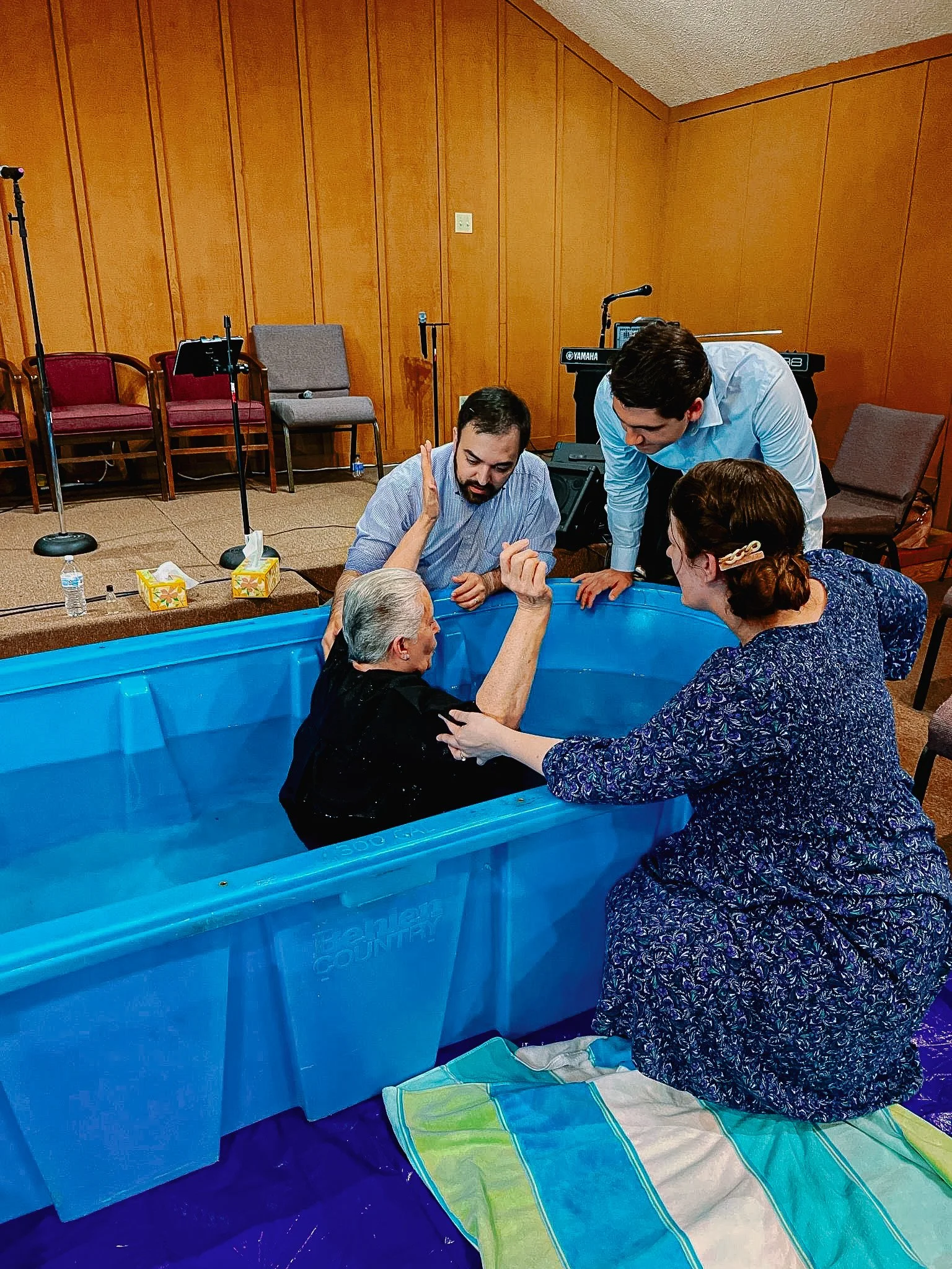 A woman being baptized in a blue plastic tub with four people supporting her during the ceremony, while musical instruments and chairs are in the background.