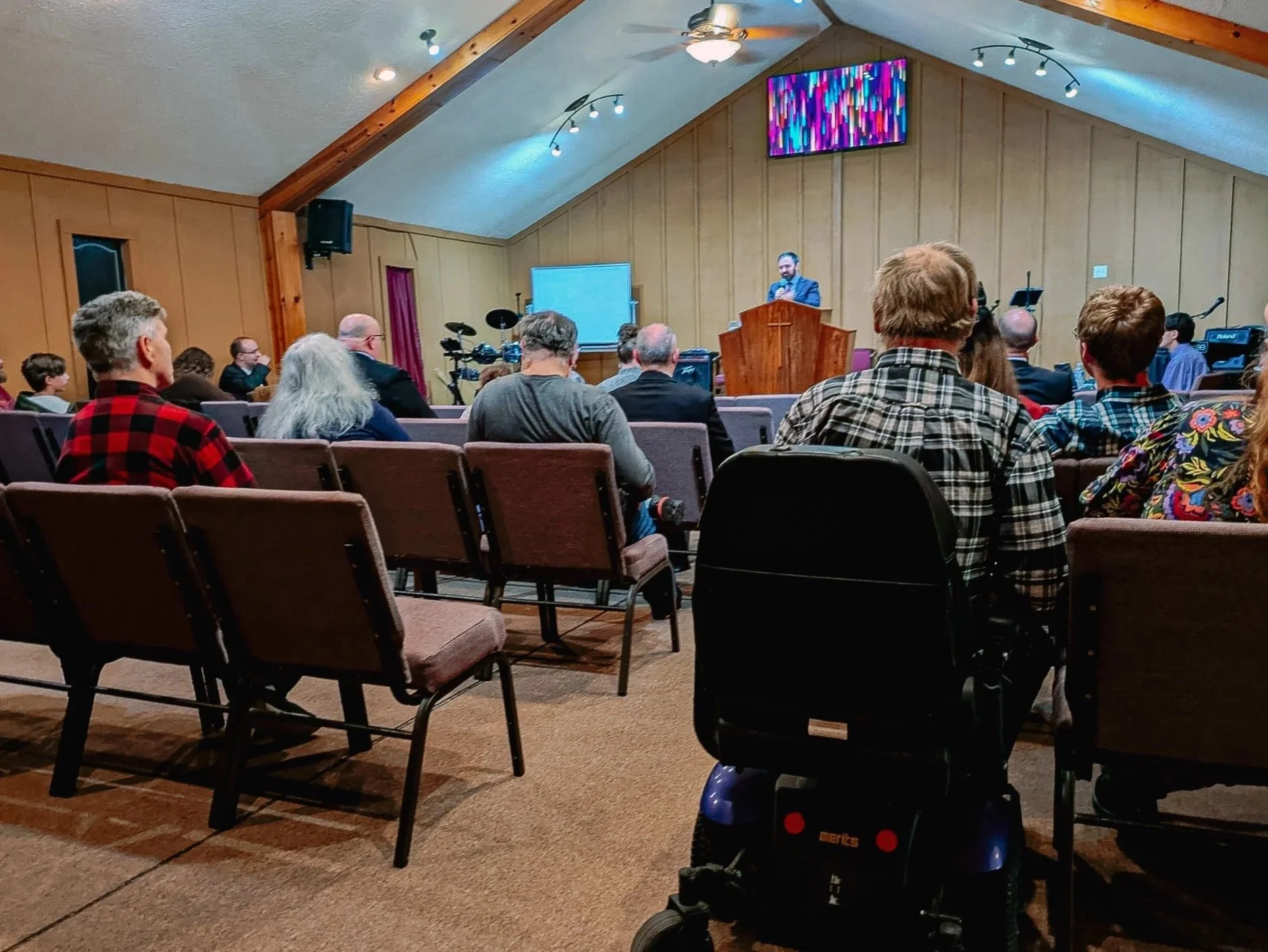 A congregation listening to a speaker at a church or event hall with wooden paneling, a large screen, and musical instruments at the front.