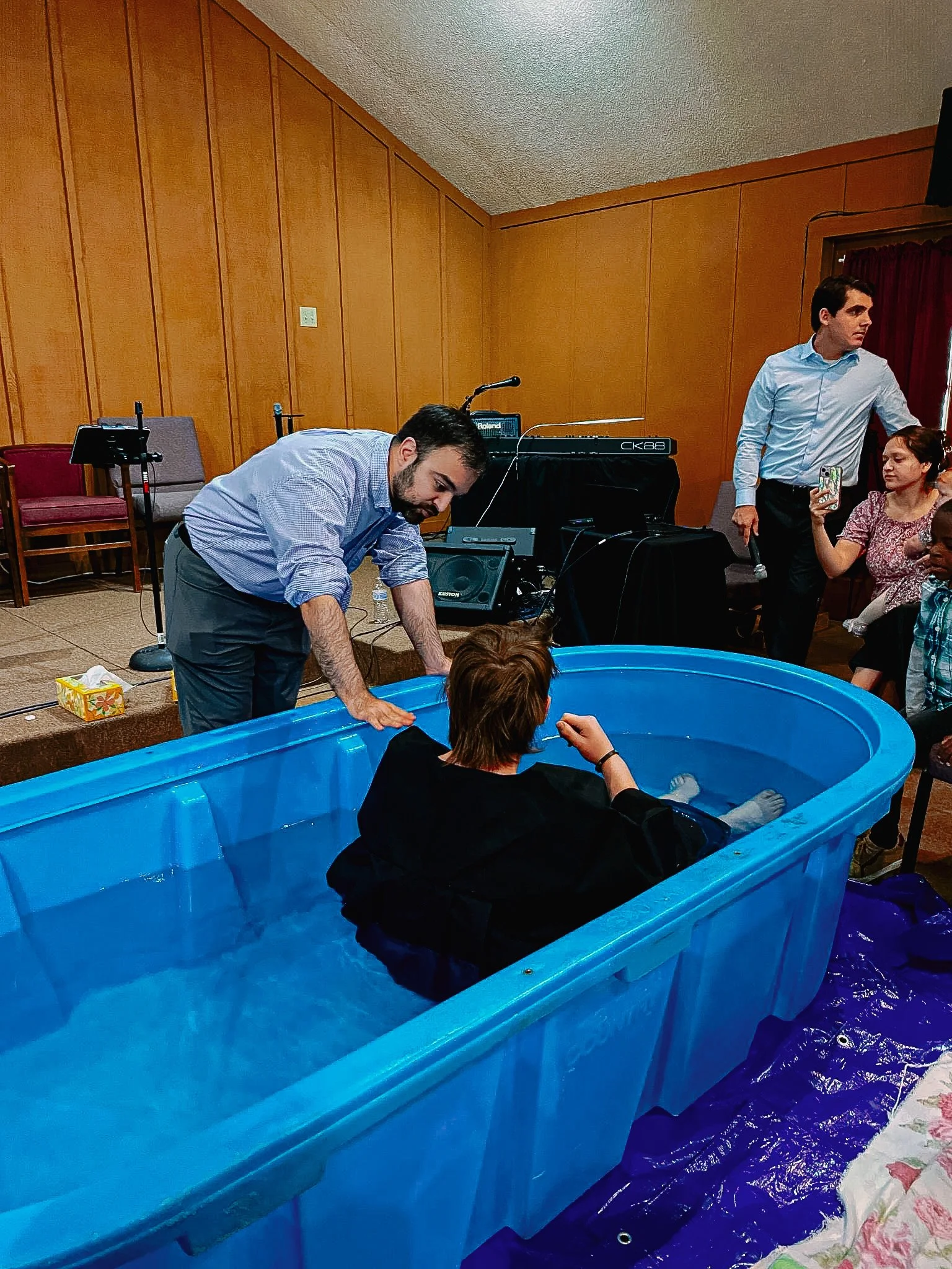 A woman is sitting in a baptismal pool while a man leans over and speaks to her. Several other people are watching, some taking photos or videos. The setting appears to be an indoor event or church service with wood-paneled walls and musical equipment in the background.