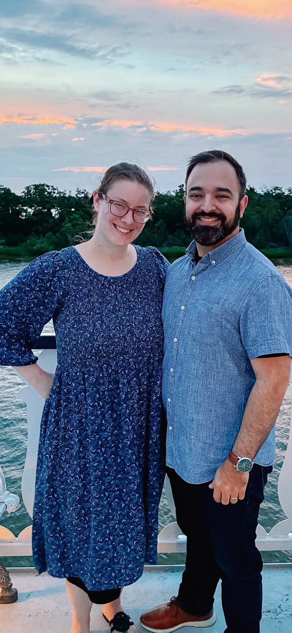 A couple posing outdoors at sunset near a body of water with trees in the background, smiling at the camera.