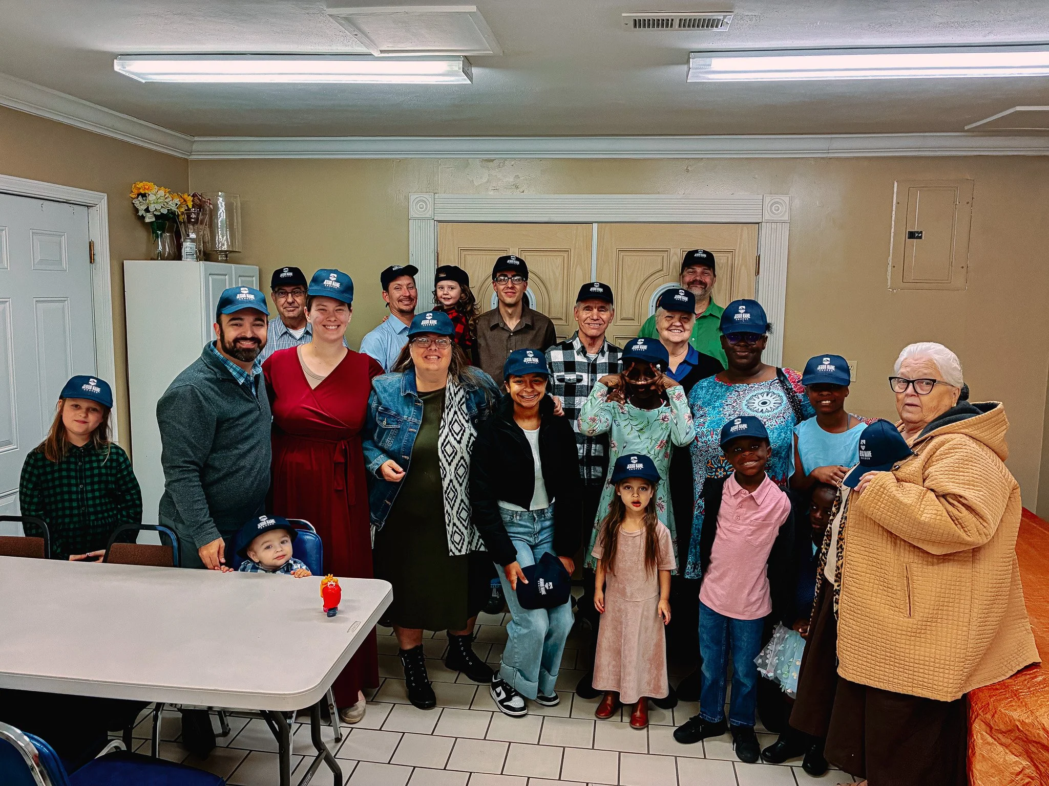 A group of people, including children and adults, wearing matching blue hats, posing inside a room with beige walls and white trim, for a group photo.