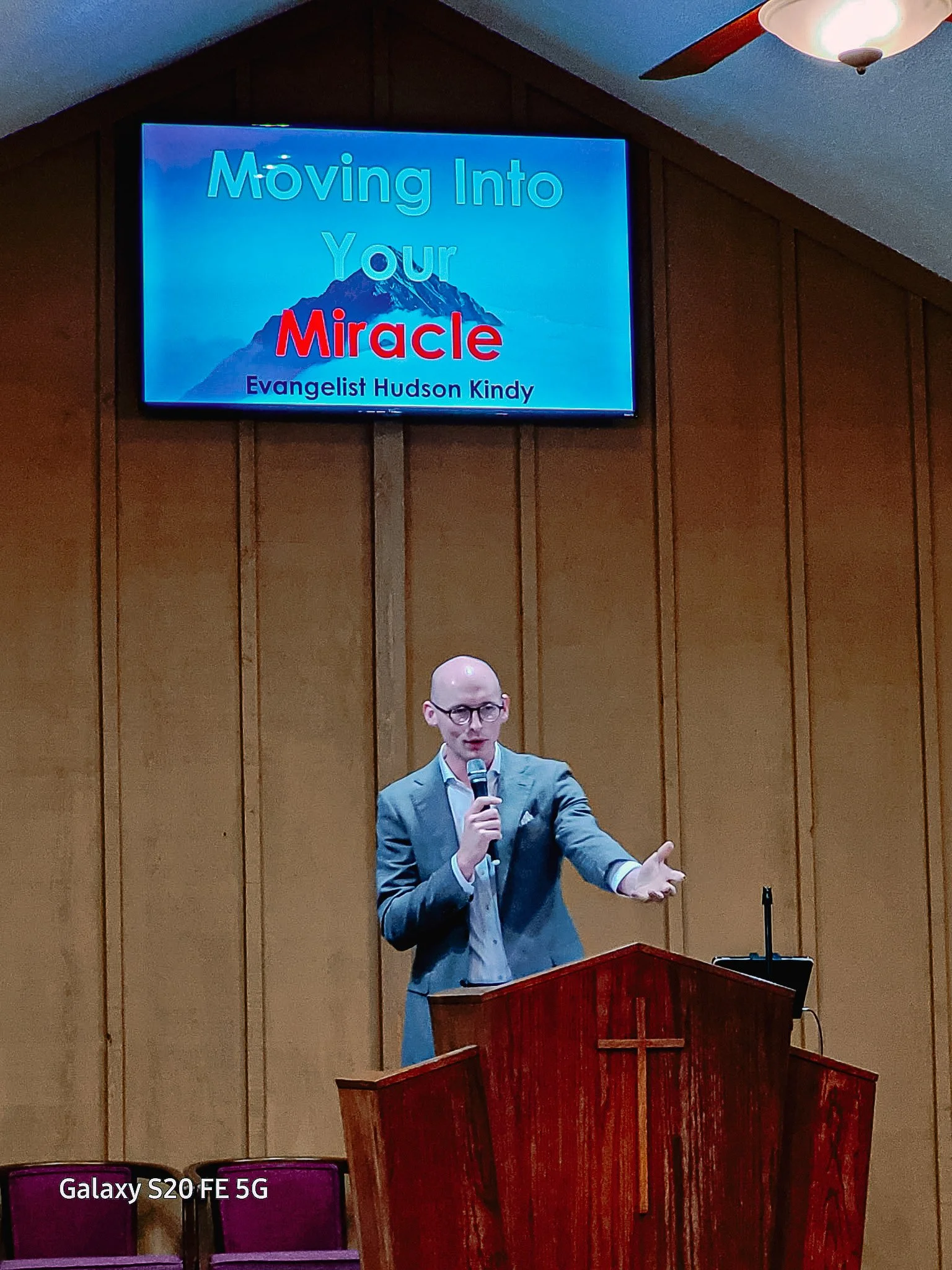 A man in a gray suit and glasses is speaking into a microphone at a church pulpit with a cross, in front of wood-paneled walls. A large screen above displays the title "Moving Into Your Miracle" and the speaker's name, Evangelist Hudson Kindy.