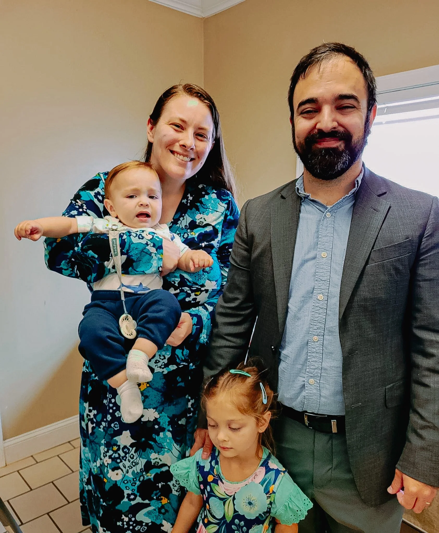 A woman holding a crying baby girl, a man in a gray suit, and a girl with red hair and hair clips standing in a room with beige walls and tile flooring.