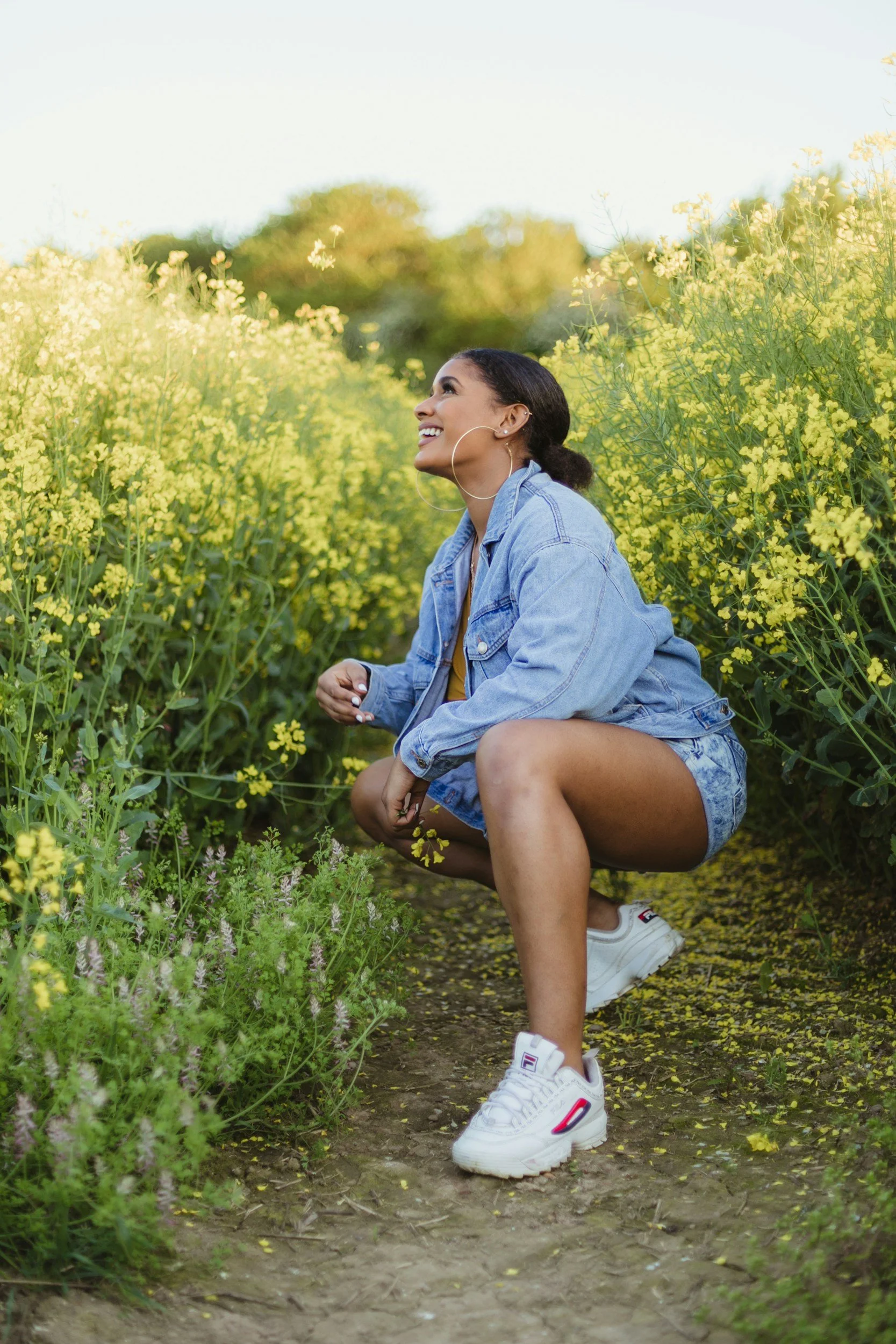 A woman crouching on a dirt path in a field of yellow flowers, smiling and looking up, wearing a denim jacket, shorts, and white sneakers.