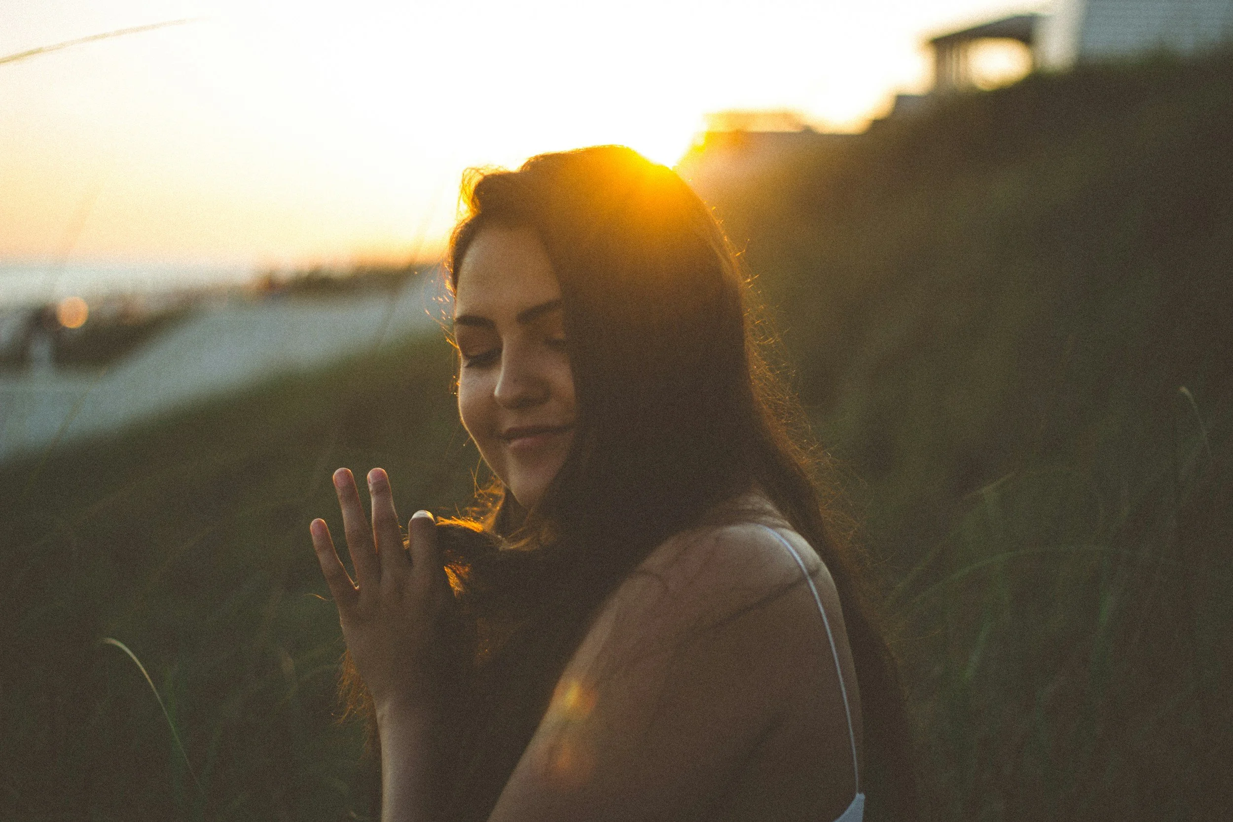 A young woman with long dark hair is outdoors at sunset, smiling softly, with her eyes closed and hands close to her face, in a peaceful pose.