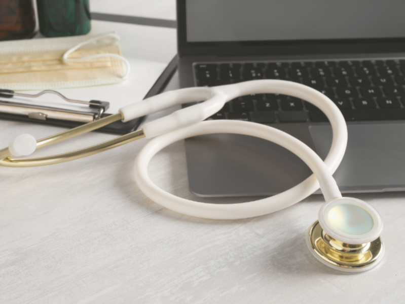 Medical stethoscope on a desk next to a laptop, with a clipboard and medical supplies in the background.