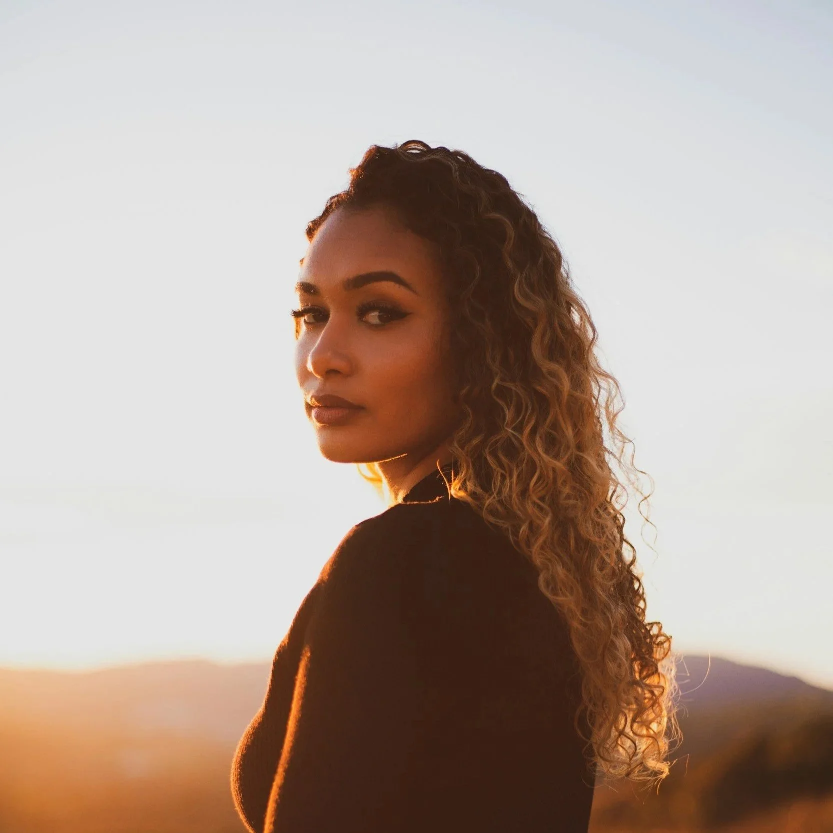 A woman with curly hair standing outdoors at sunset with a landscape in the background.
