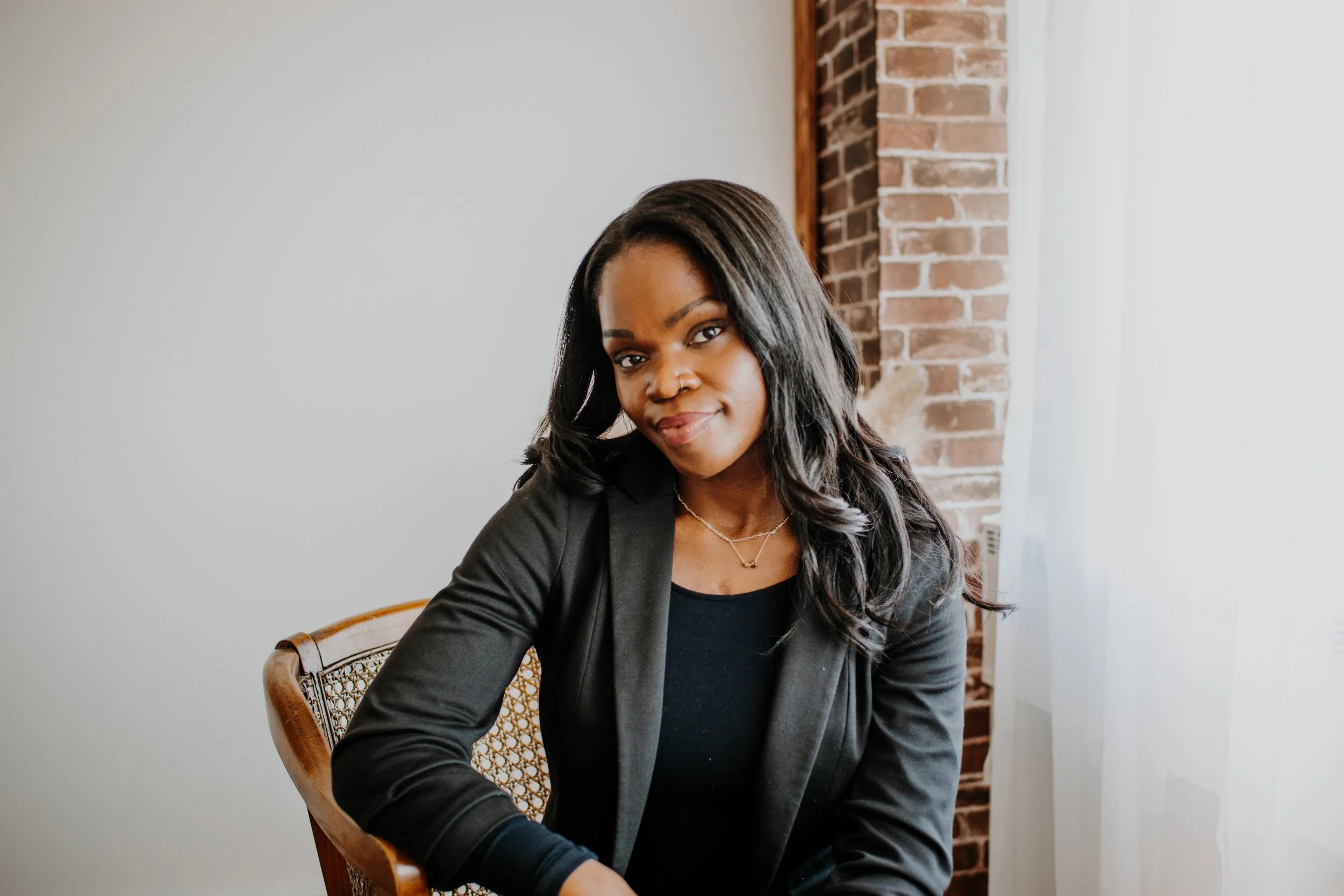 A woman with long dark curly hair and dark skin sitting indoors near a brick wall, smiling softly, wearing a black top and blazer, with a jewelry necklace.