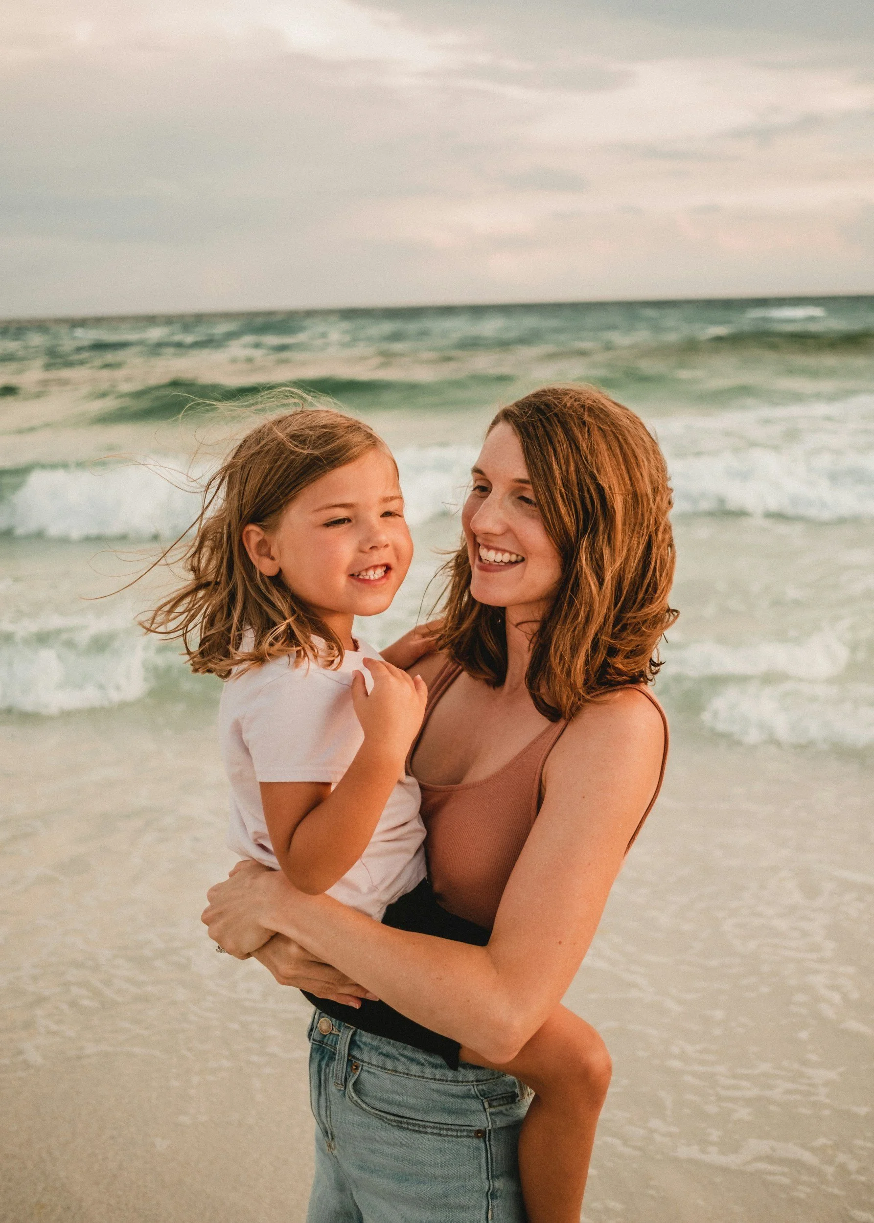 A woman with red hair holding a young girl with curly hair, both smiling, at a beach with ocean waves in the background.