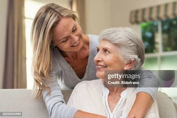 A woman and an elderly woman smiling and embracing each other indoors.