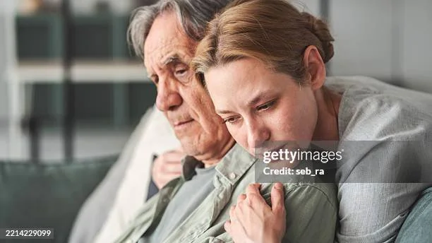An elderly man who appears sad and a daughter hugging him showing concern and love