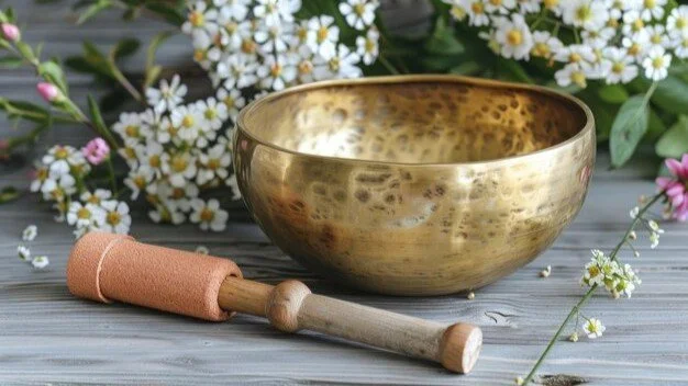 A gold singing bowl with a wooden mallet on a wooden surface, surrounded by white and pink flowers.