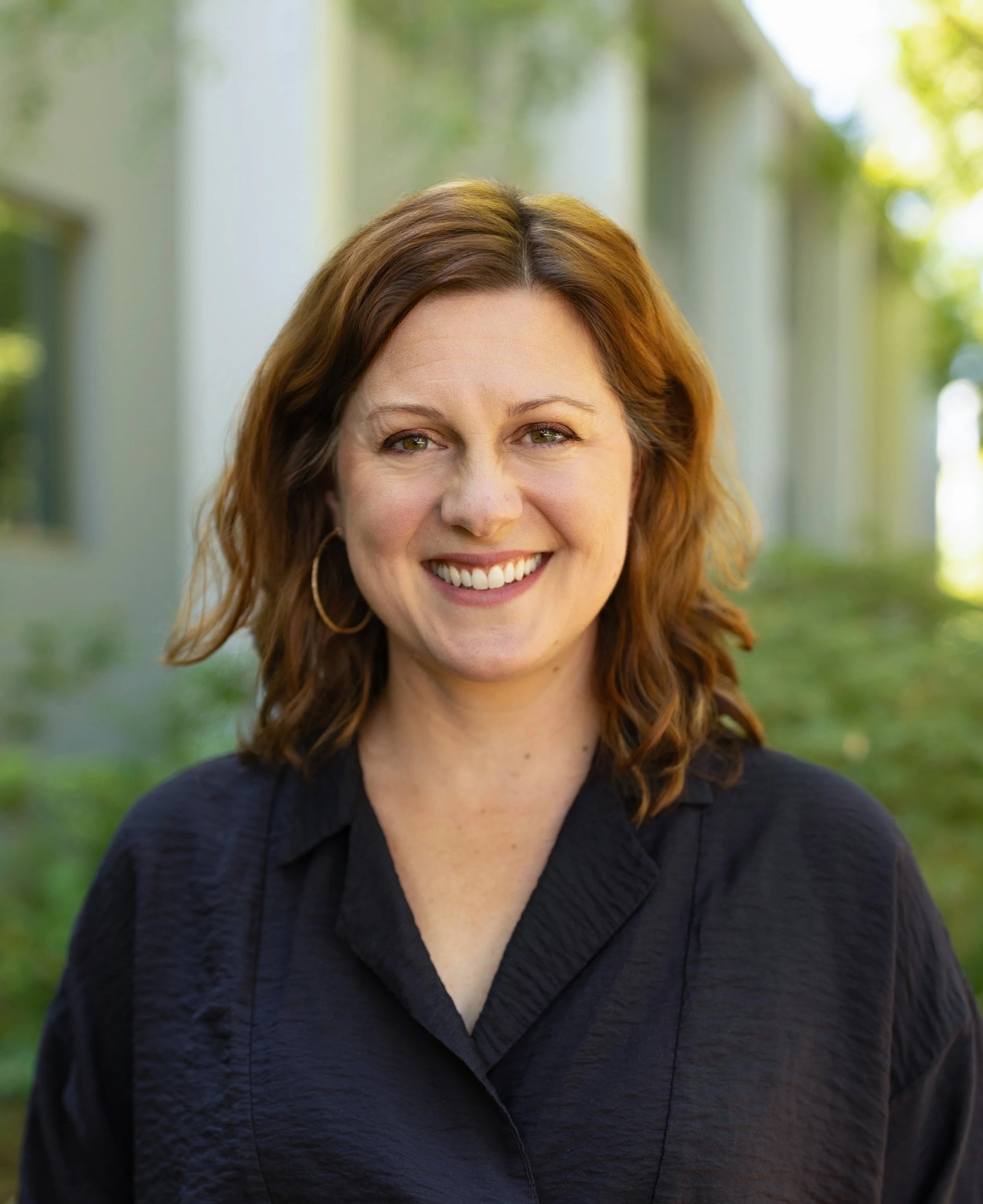 A woman with shoulder-length red hair, smiling, wearing hoop earrings and a black textured shirt outdoors with blurred greenery and a building in the background.