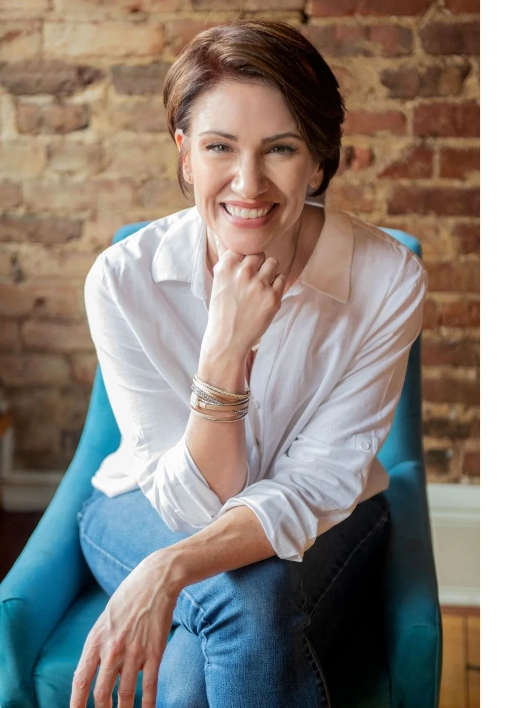 A woman with short brown hair, smiling and leaning forward with her chin resting on her hand. She is wearing a white shirt, blue jeans, and multiple bracelets. She is seated on a blue chair in front of a brick wall.