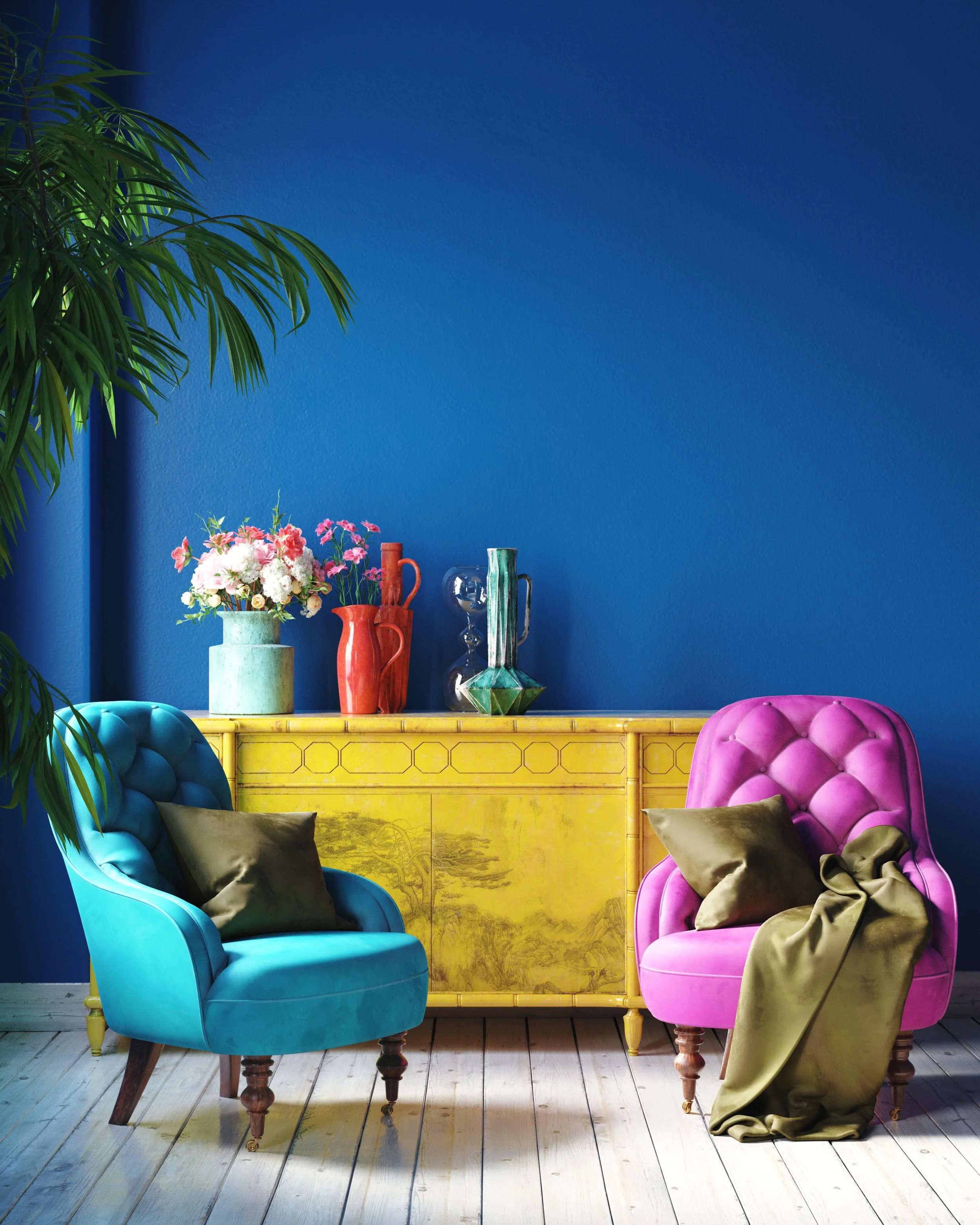 A colorful interior with a blue wall, a yellow cabinet, two velvet armchairs in blue and pink with black and brown pillows, a green and gold hat on the pink chair, and vases with flowers and decorative objects on top of the cabinet.