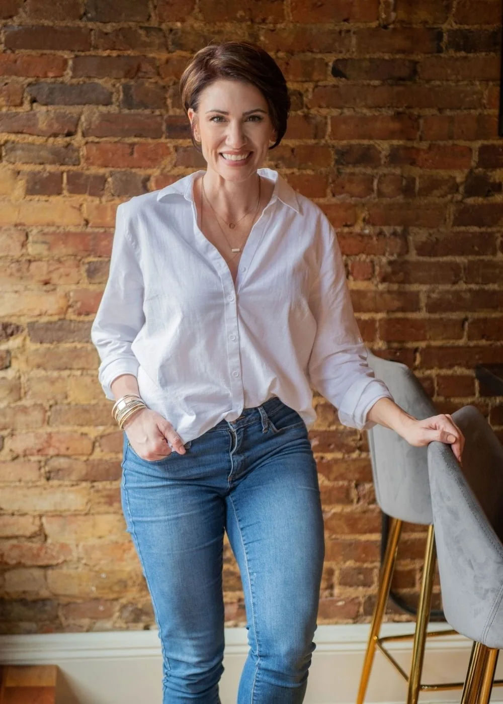 A woman with short brown hair, wearing a white button-up shirt and blue jeans, smiling and leaning on a gray barstool in front of a brick wall.