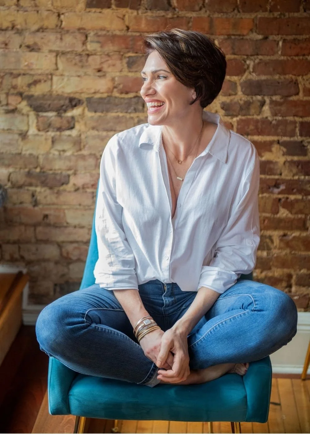 Smiling woman sitting cross-legged on a teal chair, wearing a white shirt and blue jeans, against a brick wall background.
