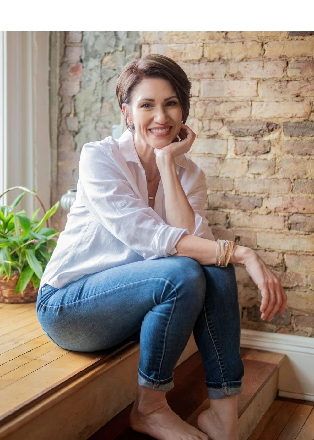 A woman with short brown hair sitting on a wooden bench near a brick wall, smiling at the camera, wearing a white shirt and blue jeans, with a potted plant nearby.