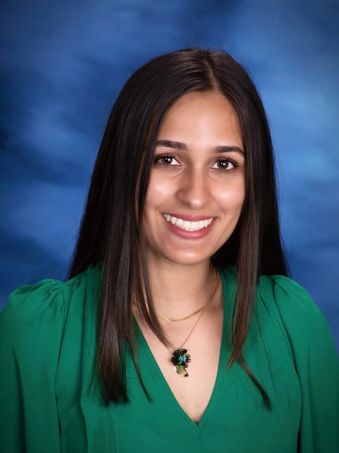 Headshot of a smiling woman with long dark hair, wearing a green top and a gold necklace with a decorative pendant, against a blue background.