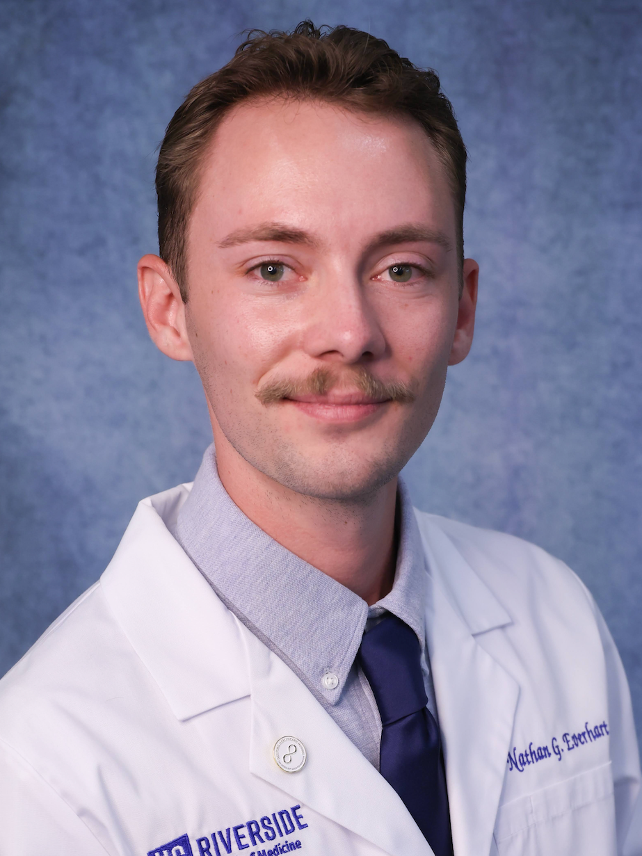 A young man with short brown hair, a mustache, and blue eyes wearing a white lab coat, a light gray button-up shirt, and a navy blue tie, standing in front of a blue textured background.