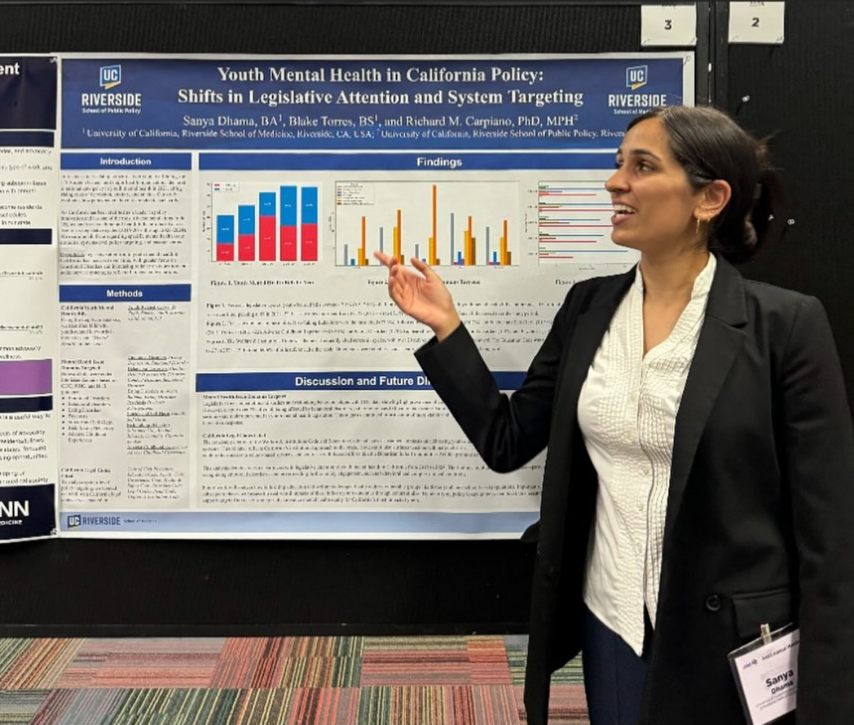 A woman in professional attire is standing next to a research poster, pointing to its findings. The poster is titled 'Youth Mental Health in California Policy: Shifts in Legislative Attention and System Targeting' and features graphs and text, with the university logo 'UC Riverside' at the top.