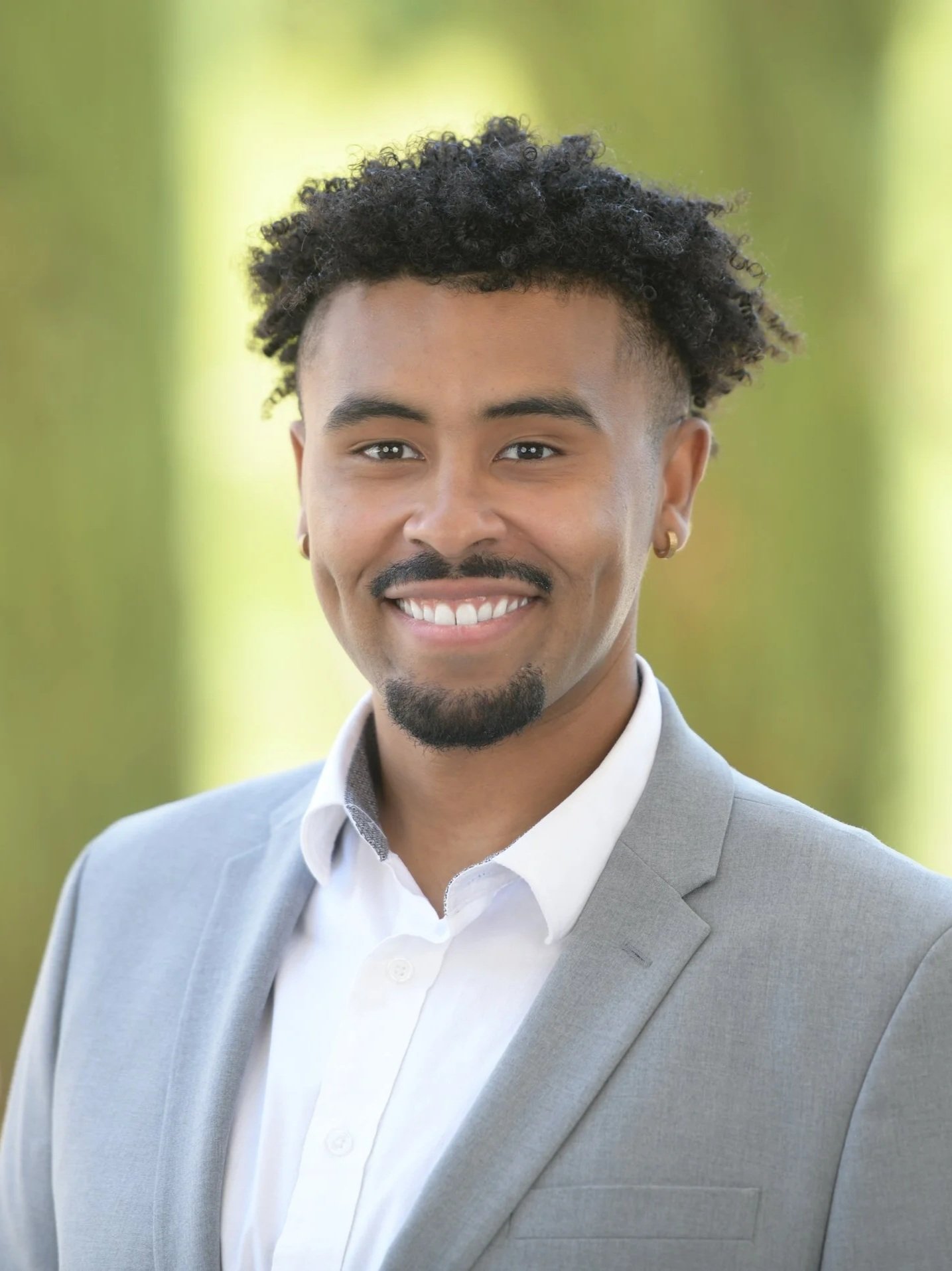 A man dressed in a light gray suit and white shirt, smiling outdoors with a blurred green background.