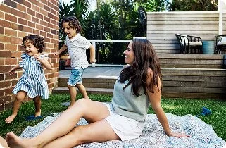 A woman and two children playing and running on a backyard lawn near a wooden fence.