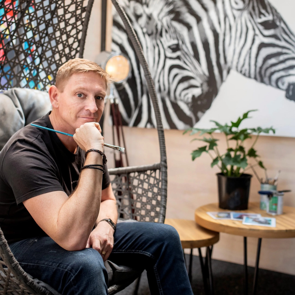 A man sitting in a hanging chair holding a paintbrush, in a room with a zebra painting, a potted plant, and photos on a small wooden table.