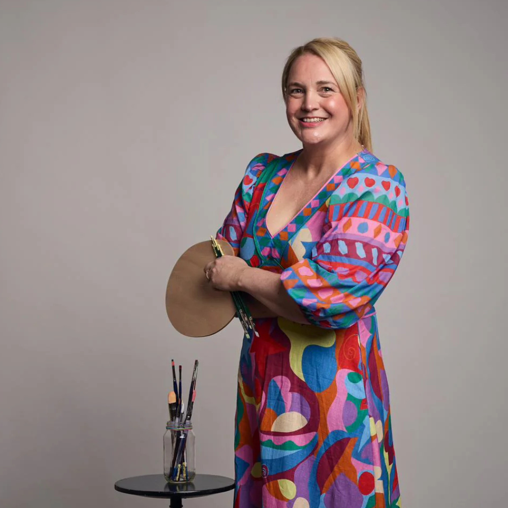 A woman smiling and holding a small art palette with brushes, standing next to a table with paintbrushes in a jar, wearing a colorful, patterned dress with bright geometric and abstract shapes.