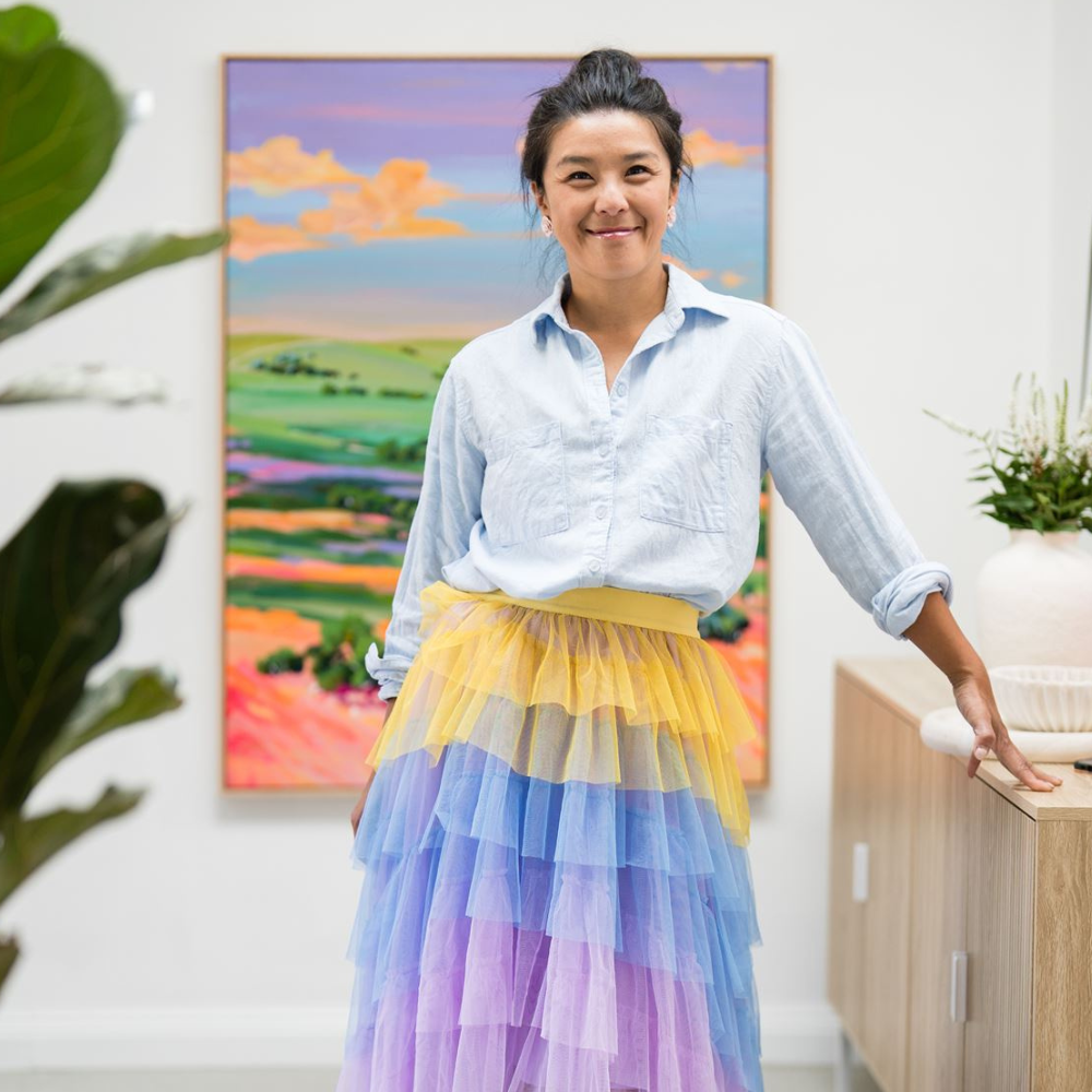 A woman with dark hair in a bun, smiling, wearing a light blue button-up shirt and a colorful tiered tulle skirt in yellow, blue, and purple, standing in front of an abstract landscape painting with green fields and pink skies in an art gallery or studio.