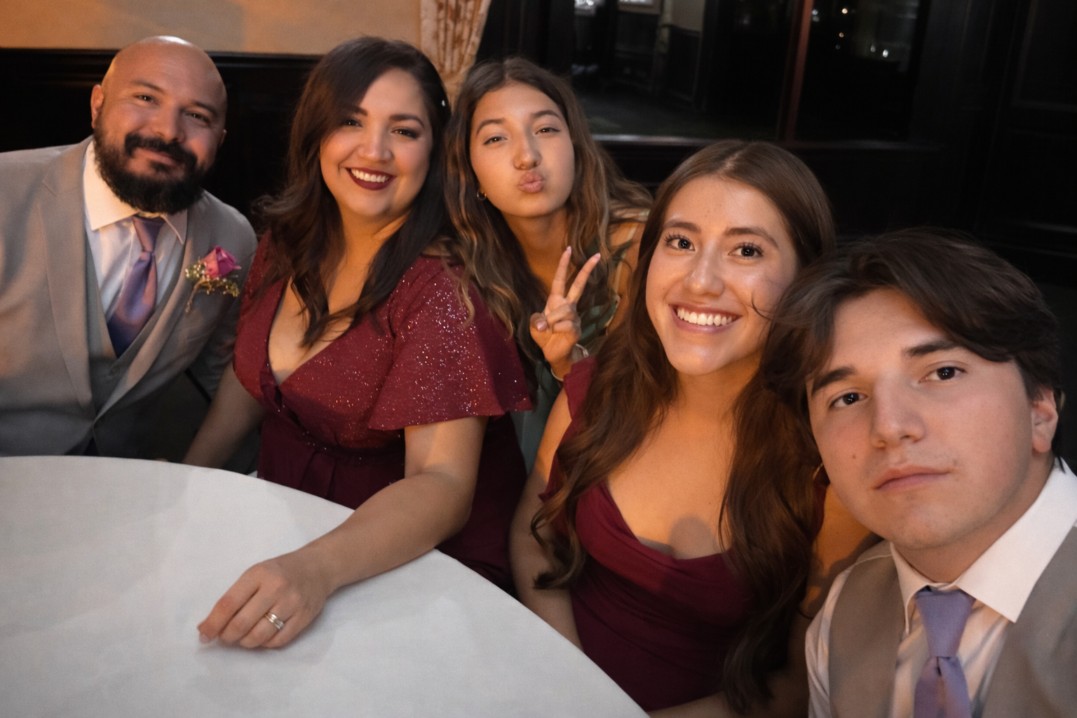 Group of six people smiling and taking a selfie at a dinner event, seated around a table with a white tablecloth, in an indoor venue.