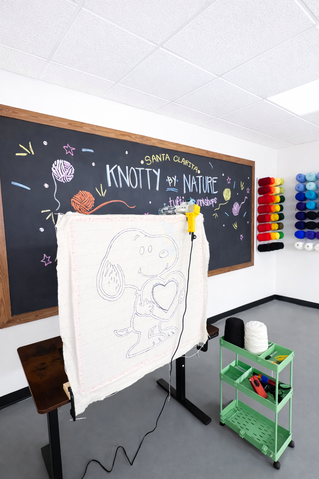 A knitting workshop room with a blackboard that reads "Knotty by Nature," and a large fabric with a Snoopy drawing on an easel. There are yarns in various colors on the right, and a green cart with sewing supplies, along with a black and a white rolled-up fabric.