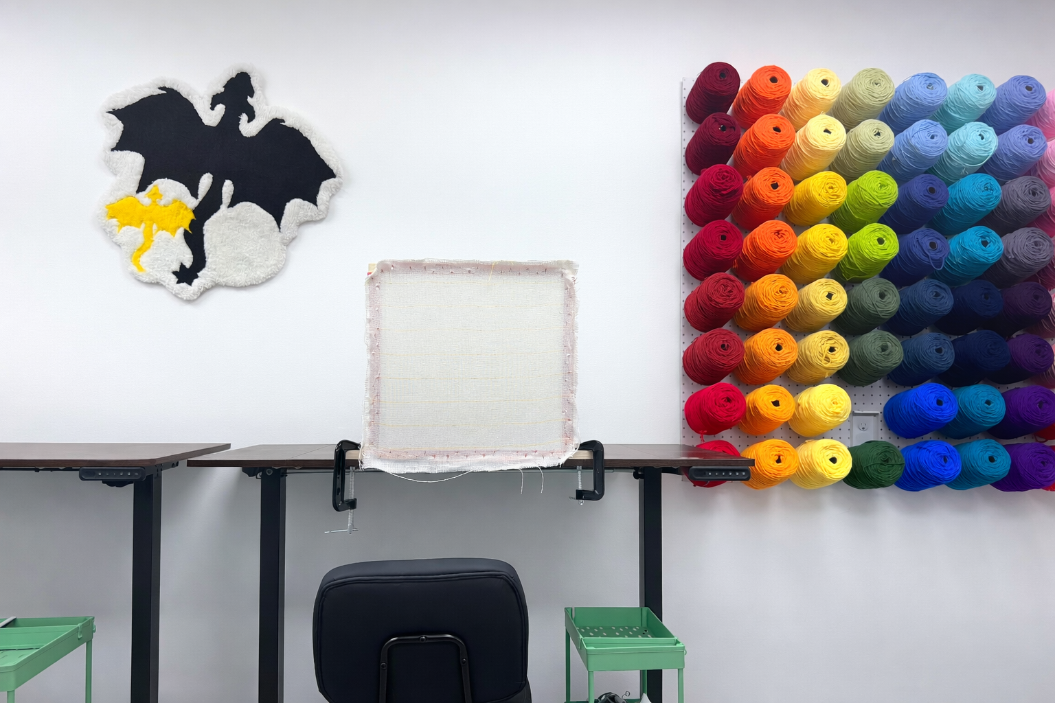 A wall with a colorful display of yarn spools organized in a grid pattern on the right, a black and yellow dragon-themed rug hanging on the left, a sewing or craft table in the center with a clamp board, and a black chair with green storage baskets underneath.