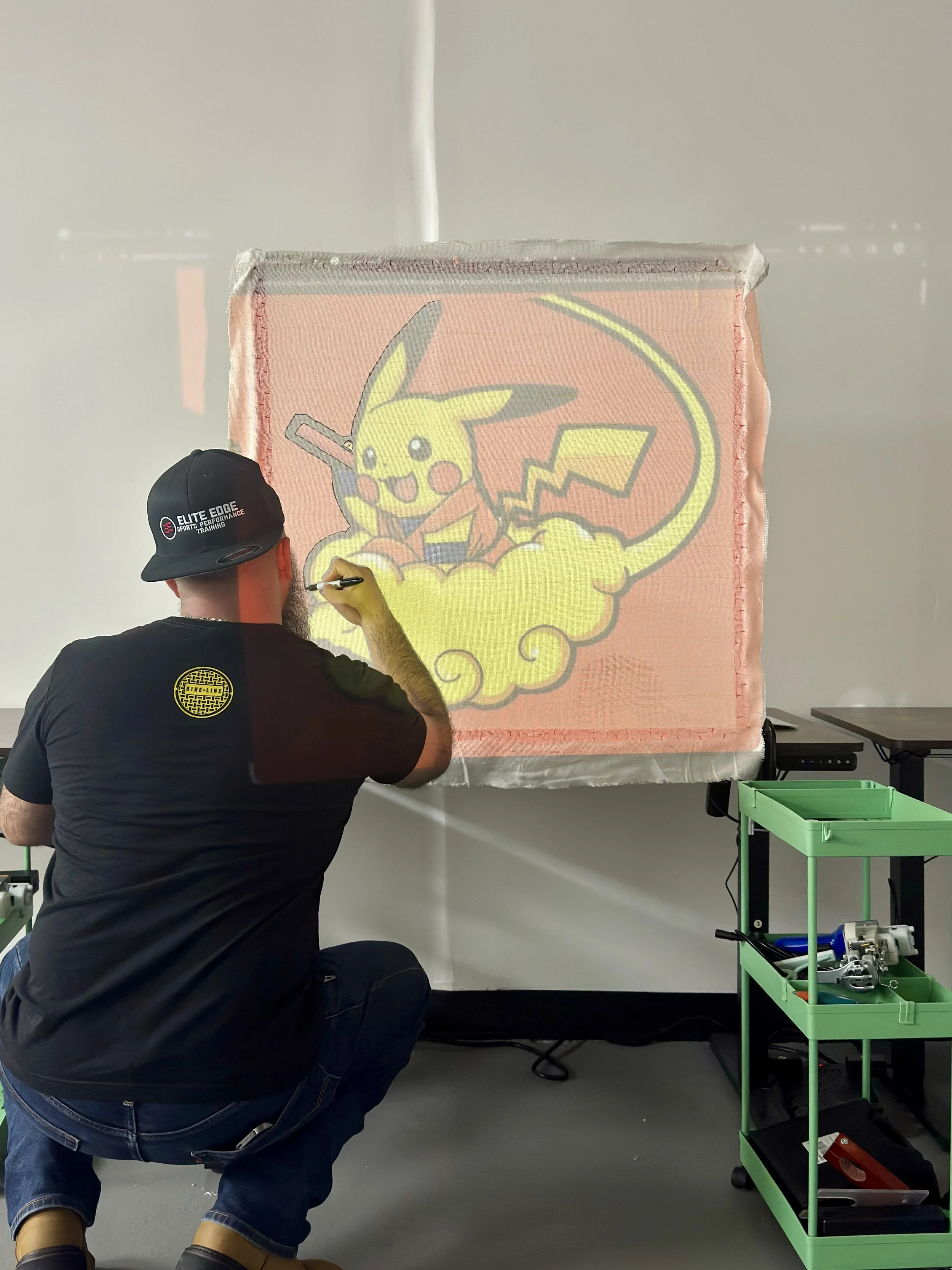 An artist is drawing a large, colorful image of Pikachu on a padded, pink surface in a workshop or studio. The artist is wearing a black shirt and a black cap that says 'Elite Edge Sport Performance Training'.