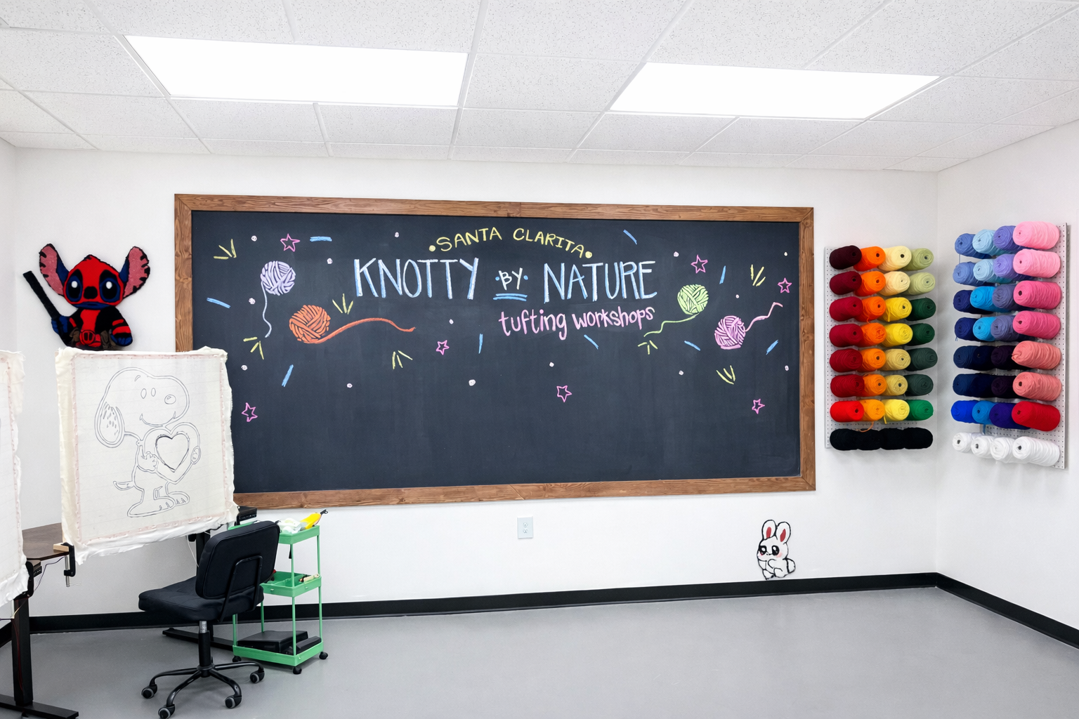 Interior of a knitting workshop room with a large black chalkboard on the wall, decorated with colorful yarn drawings and the words "Knotty by Nature" and "Santa Clarita tufting workshops." To the left, a cartoon character of Snoopy holding a heart drawing, and a stuffed toy of Stitch from Lilo & Stitch. On the right, shelves filled with colorful spools of yarn arranged by color.