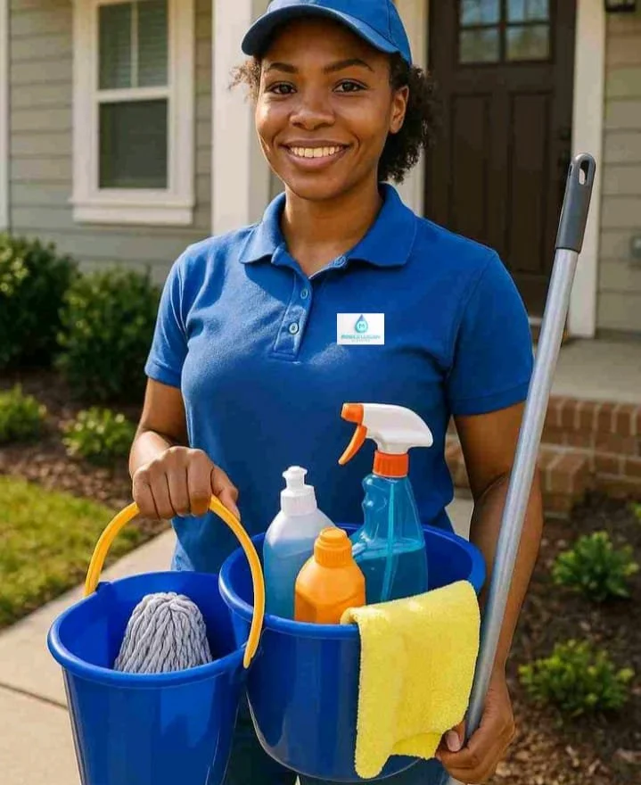 A smiling woman in a blue uniform and cap holding a blue bucket with cleaning supplies, including spray bottles, a scrub brush, and a yellow cloth, standing outside a house.