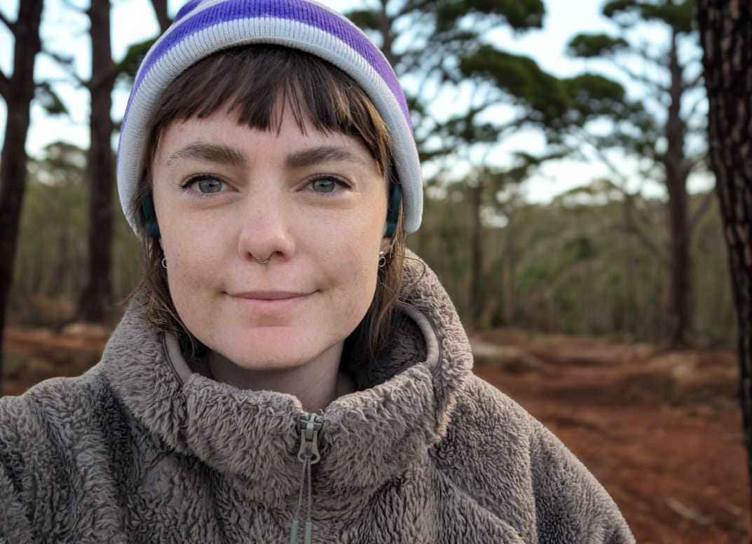 Young fair skinned woman with a short brown fringe in outdoor setting with trees, wearing a grey fleece jacket, a purple and white beanie, and earbuds.