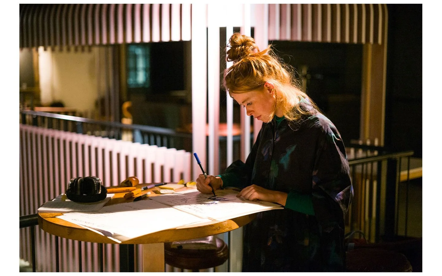 A woman with red hair tied in a bun, wearing a dark patterned jacket, is drawing or writing on paper at a wooden table. The table has color markers and a pair of headphones. The background features a modern interior with wooden bars and a dimly lit ambiance.