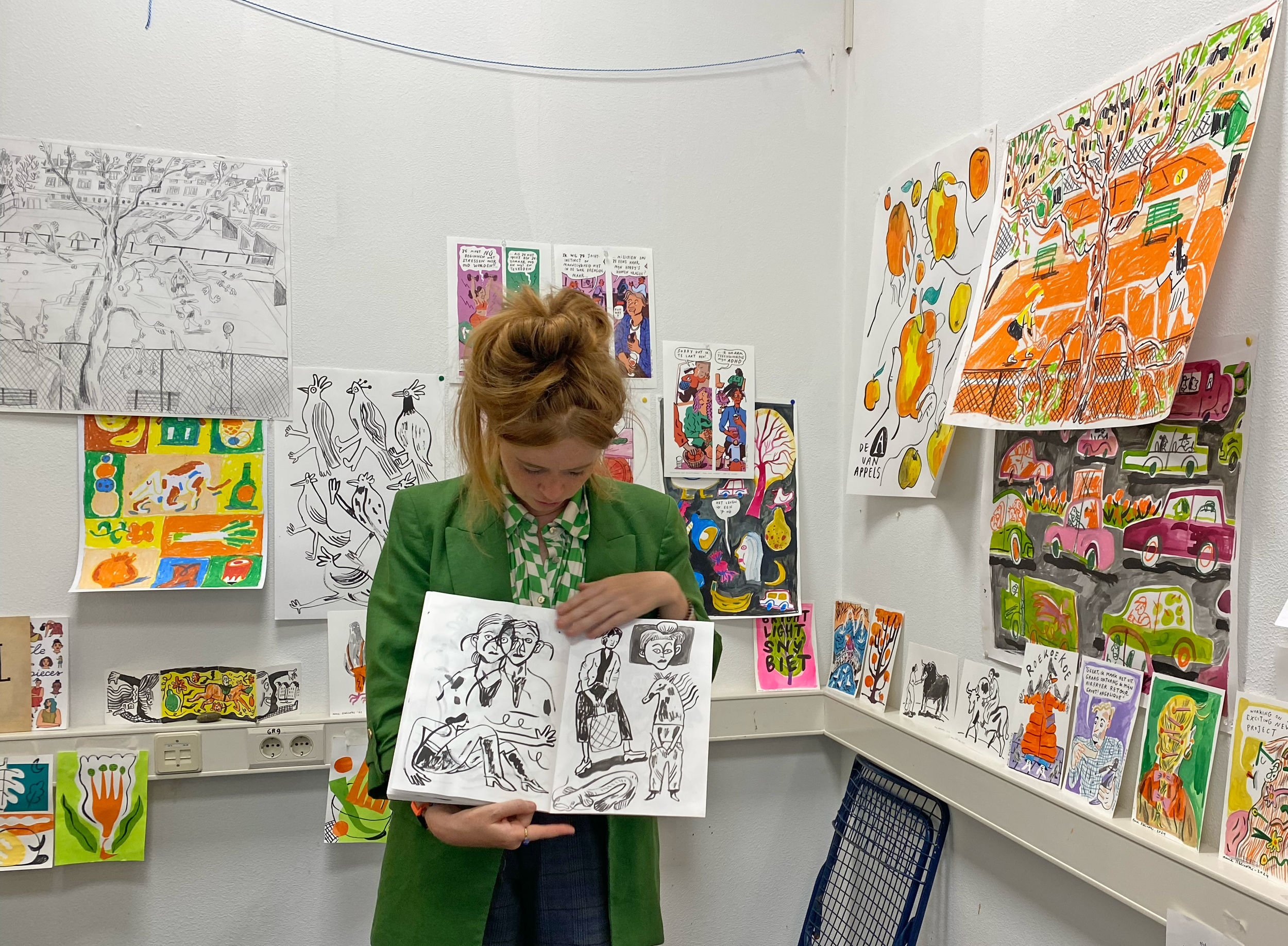 A woman in a green blazer and checkered shirt holding a sketchbook with black line drawings inside an art gallery with various colorful artworks on the walls.