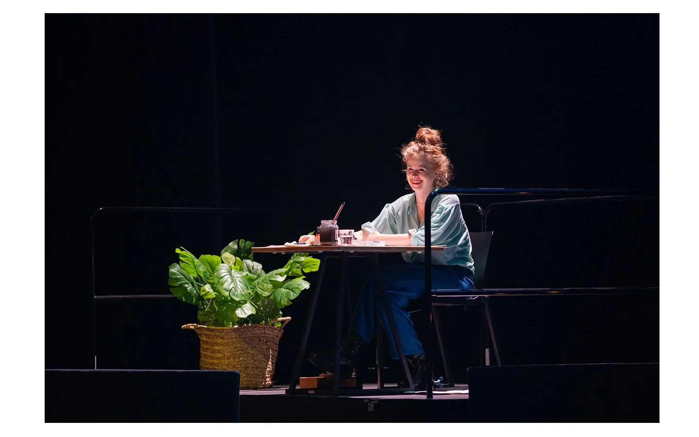 A woman with blonde hair tied in a bun, sitting at a table on stage, smiling and looking to her left. The table has a small plant, a jar of dark substance, and some papers. There is a large green potted plant in front of her, and she is wearing a light-colored top and dark pants. The background is black.